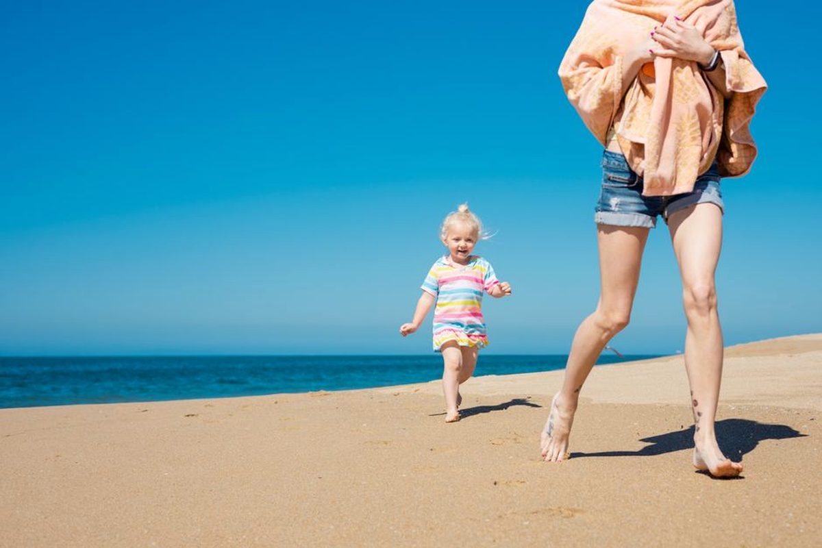 A toddler running along a sunny beach with a parent