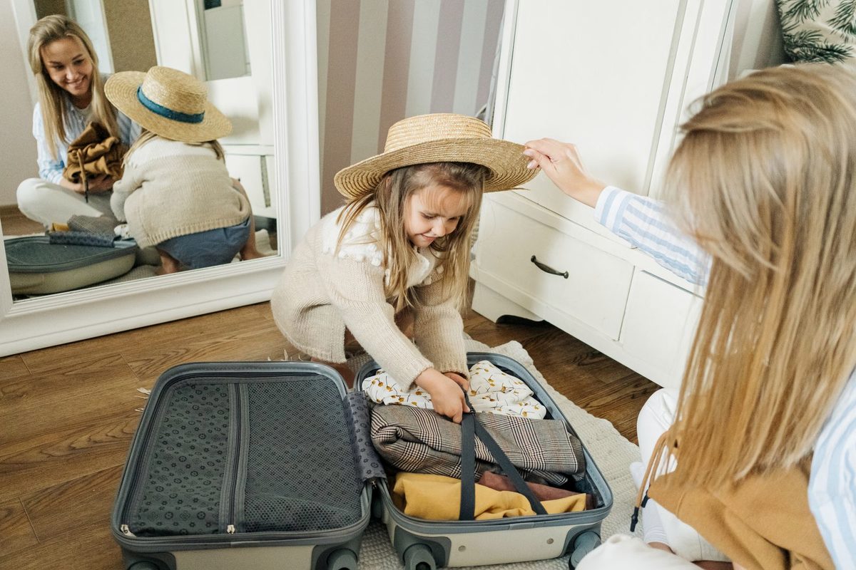 A mother and her young daughter packing a suitcase together at home, the child trying on a straw sun hat.