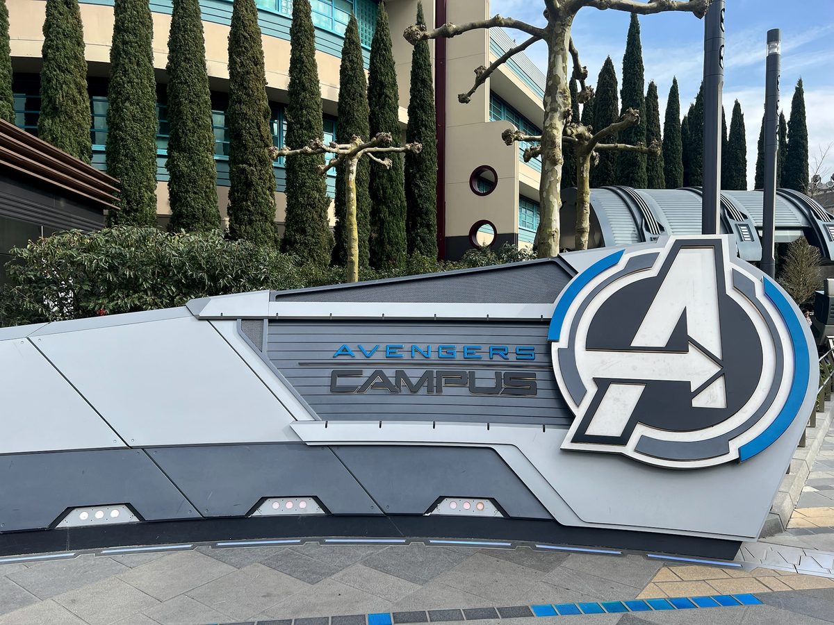The metal Avengers Campus entrance sign with the Avengers A logo at Disney Adventure World, cypress trees and blue sky behind