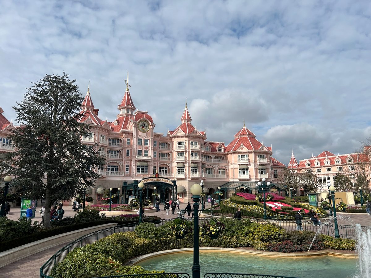 The pink turreted Disneyland Hotel rising above the flowerbeds and fountains at the end of the Disney Village walkway, under a bright partly cloudy sky