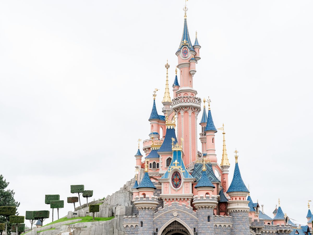 Sleeping Beauty Castle seen from below against a pale sky, its pink turrets and blue spires rising above a rocky outcrop with clipped topiary trees