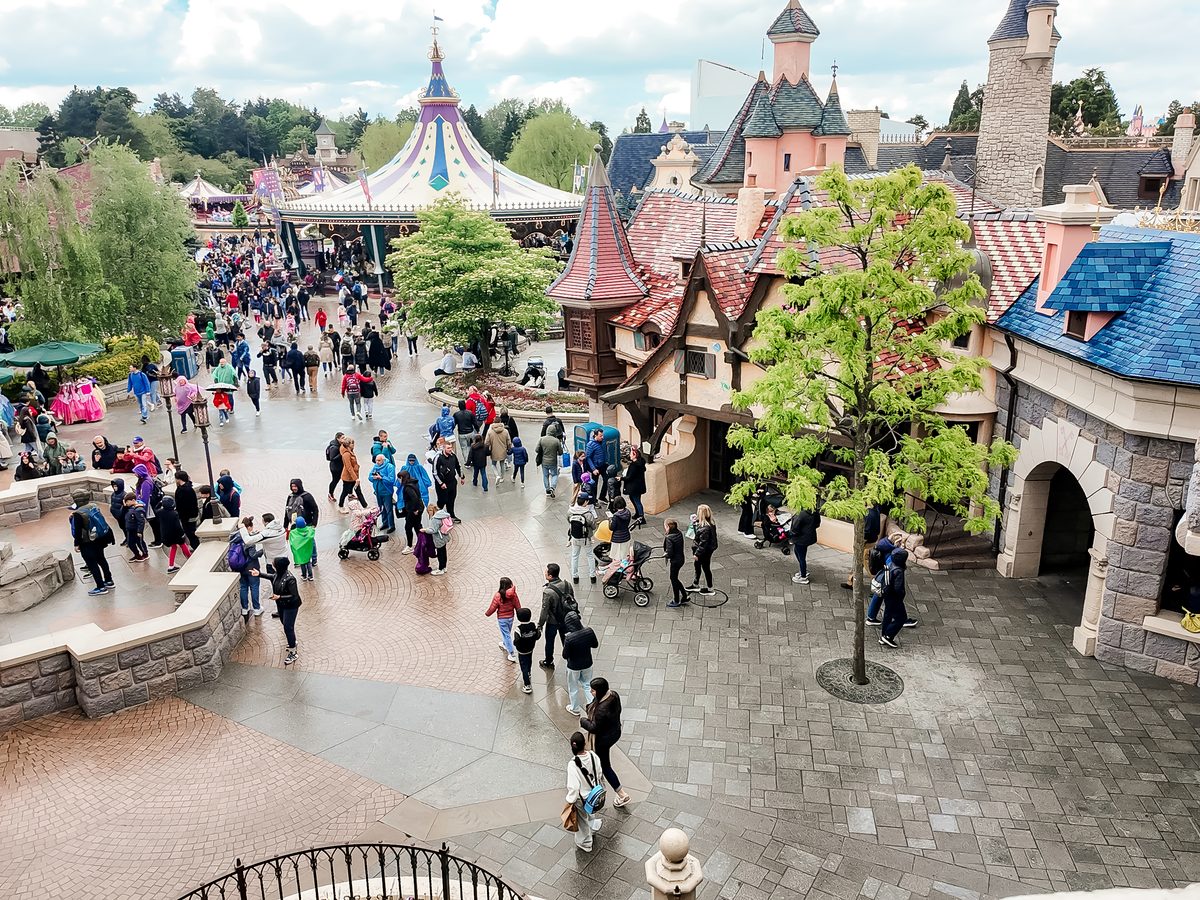 An aerial view over Fantasyland at Disneyland Park, families and pushchairs crossing the paths below the castle turrets and a striped entertainment tent