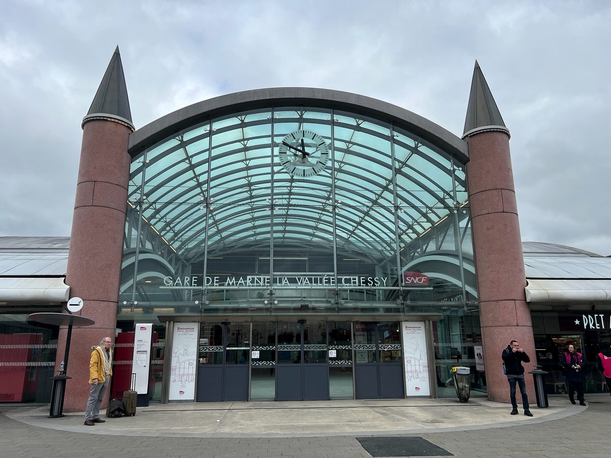 The glass arch and pink granite towers of Marne-la-Vallée-Chessy station, with a clock at the top of the glazing