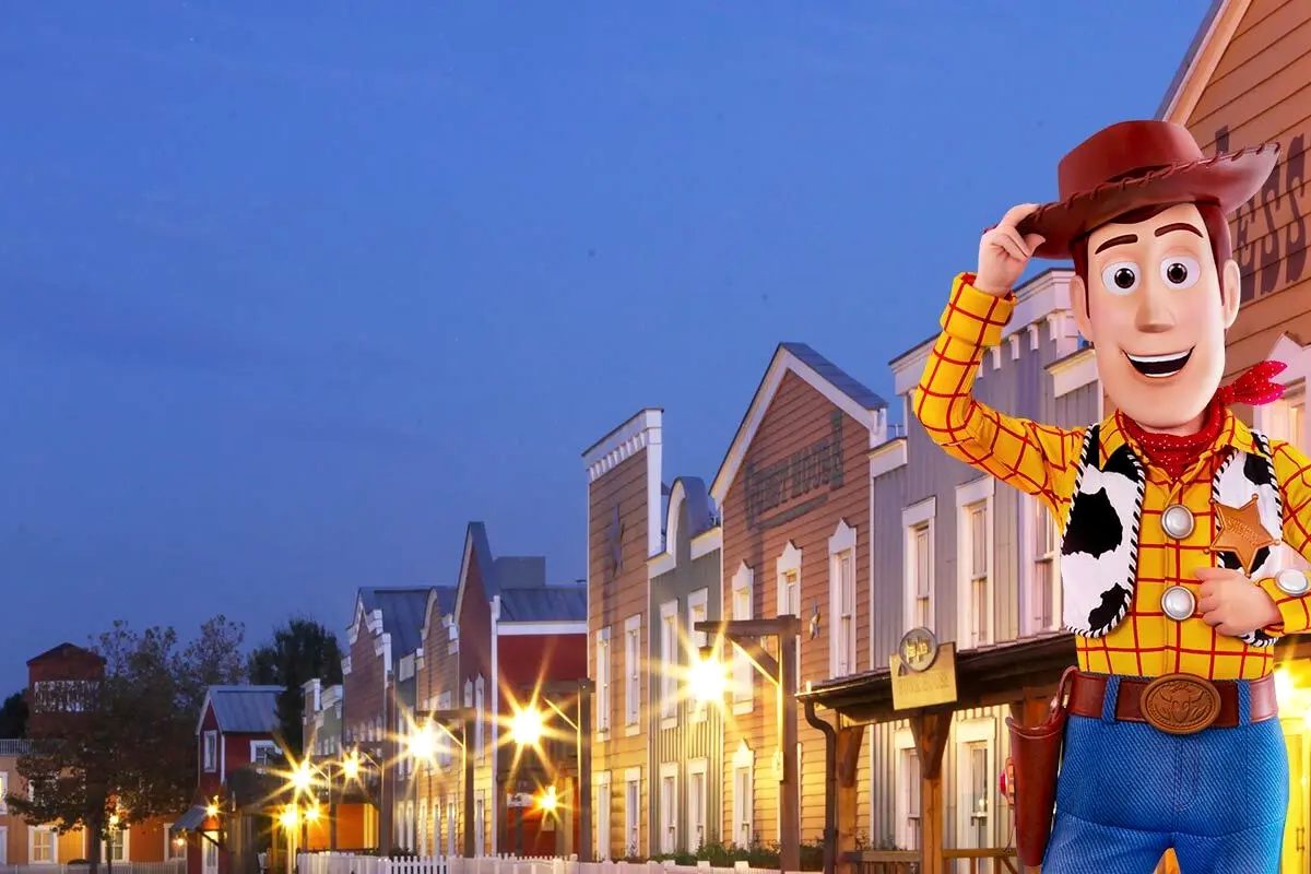 The frontier main street at Disney's Hotel Cheyenne at dusk, timber-frame Wild West buildings under lit street lamps with Woody tipping his hat in the foreground