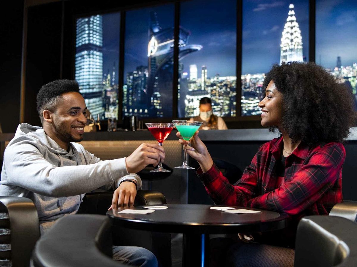 Two guests raising cocktails at the Skyline Bar, in front of window screens showing a night-time Manhattan skyline