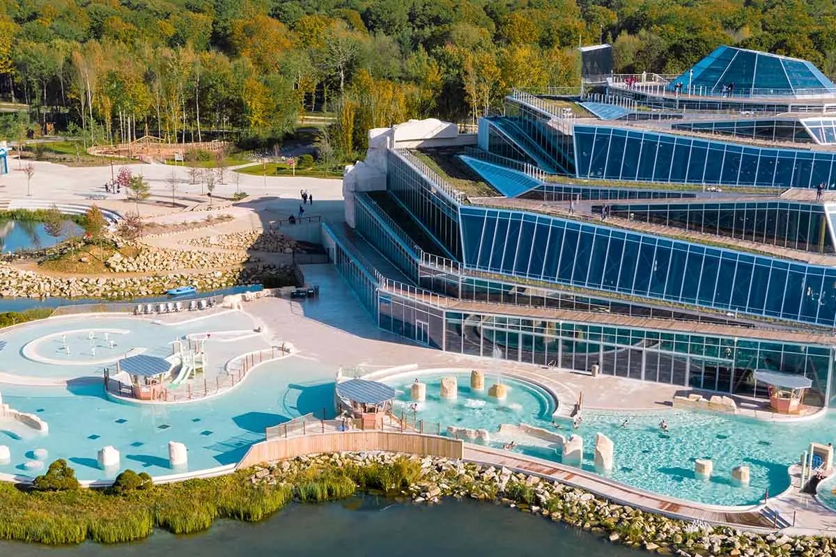 An aerial view of Villages Nature Paris, the glass Aqualagon building beside its outdoor geothermal lagoon pools, ringed by autumn woodland