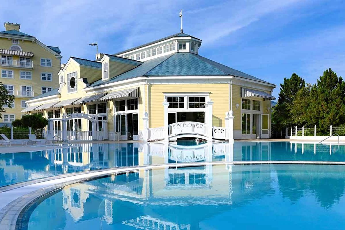 The outdoor pool at Disney Newport Bay Club, a wide blue pool with a small white footbridge and the yellow-clapboard, blue-roofed pool house behind
