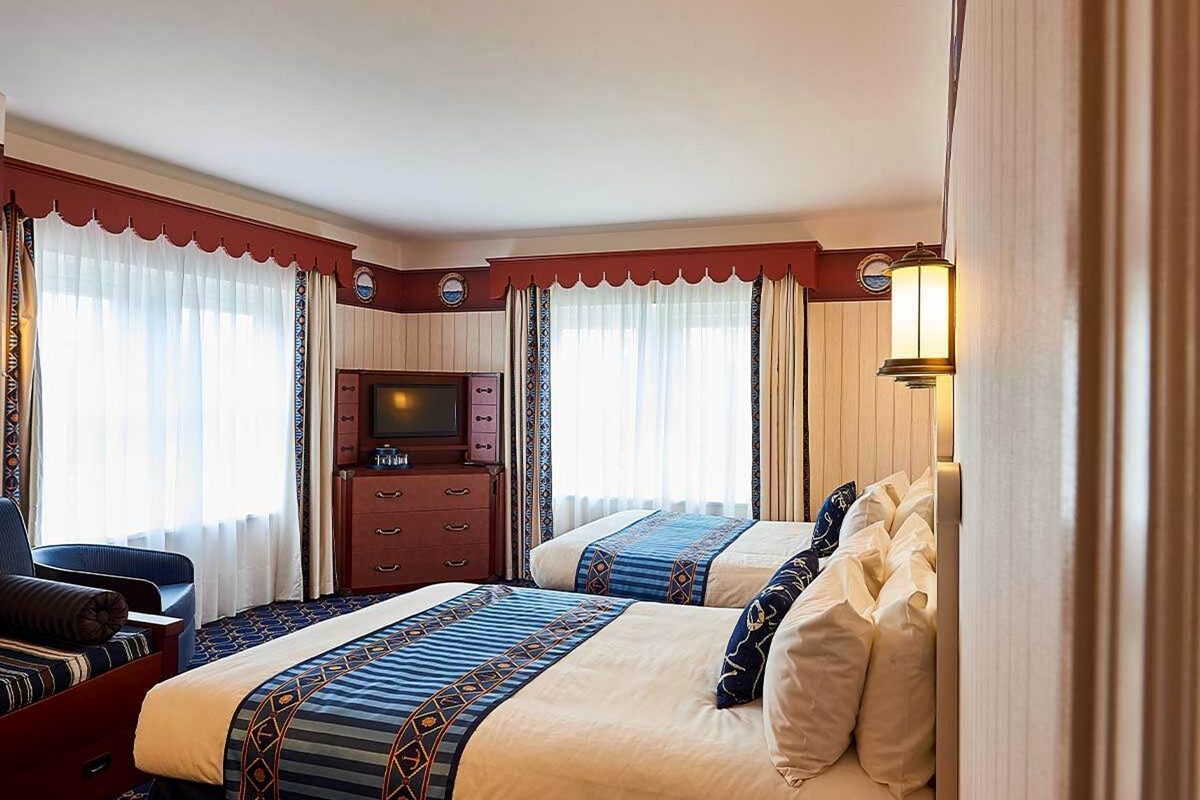 A Superior Room at Disney Newport Bay Club with two double beds, navy-and-cream striped runners, a red pelmet and a trunk-styled dresser holding the television