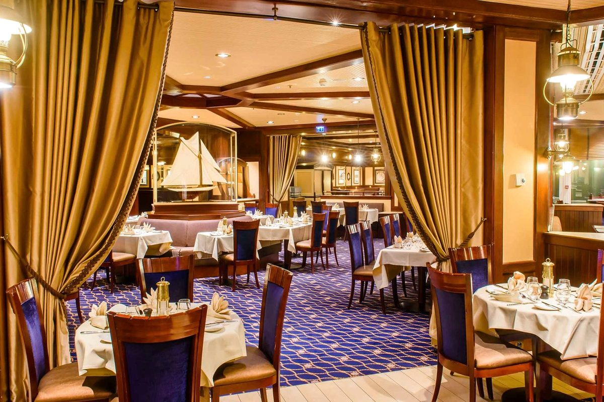 The Yacht Club dining room at Disney Newport Bay Club, gold curtains, a deep-blue rope-patterned carpet, brass lantern lamps and a model sailing ship in a case, tables set with white linen