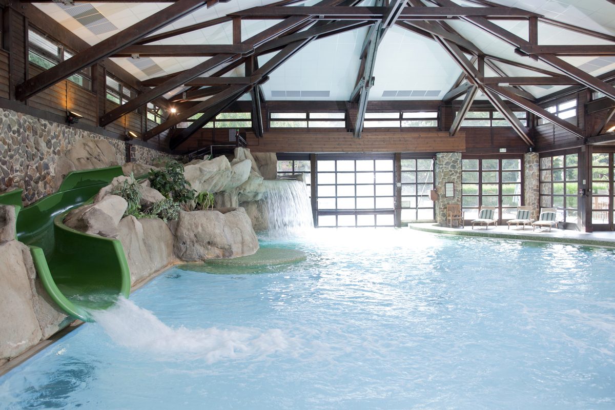 The Quarry Pool at Disney Sequoia Lodge, an indoor pool beneath timber roof beams with a green waterslide, a rockwork waterfall and loungers along the far side