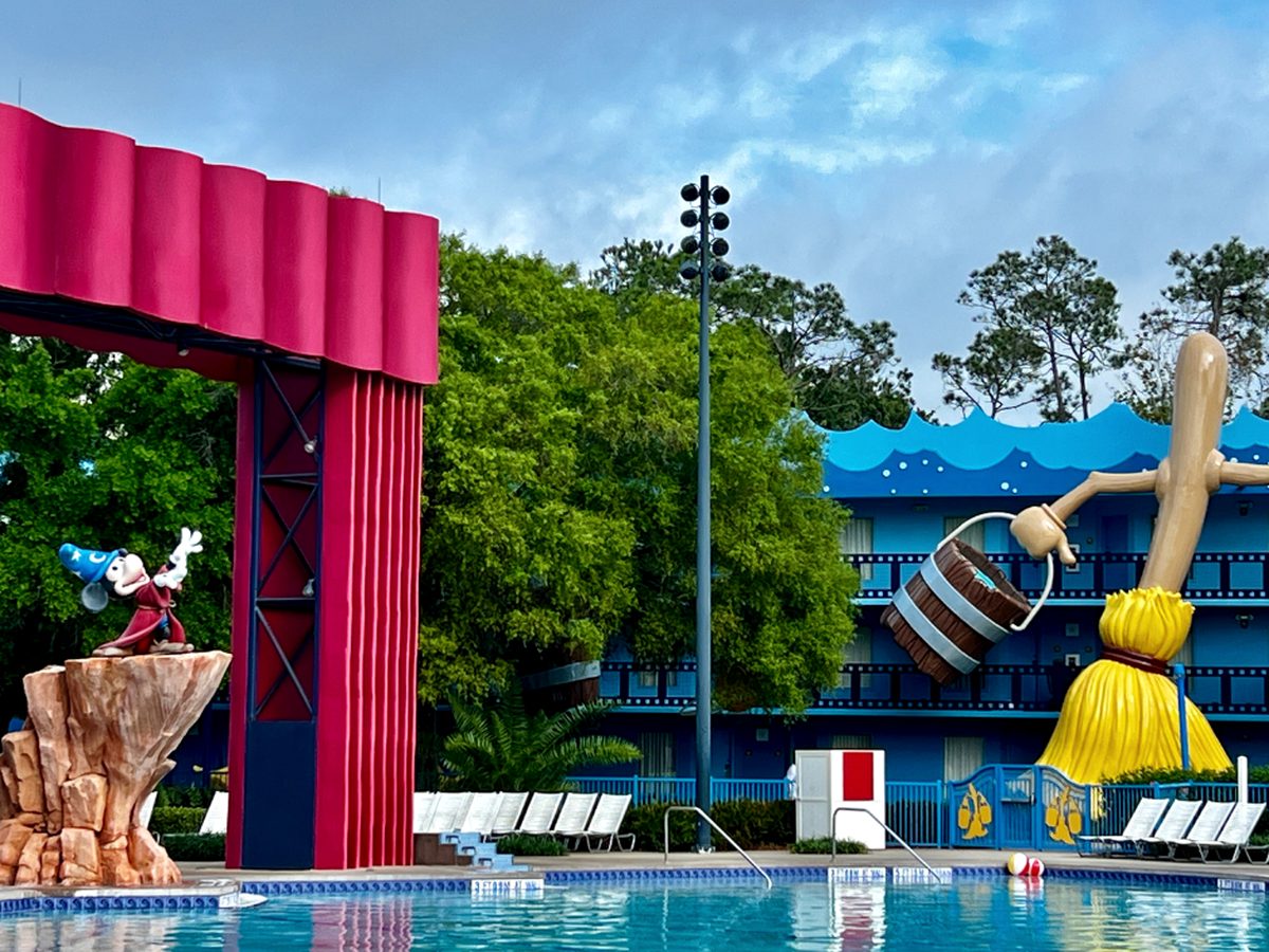 The Fantasia Pool at Disney All-Star Movies, with the Sorcerer Mickey fountain on a rock beside a giant red stage curtain and a bucket-carrying broomstick at the far side