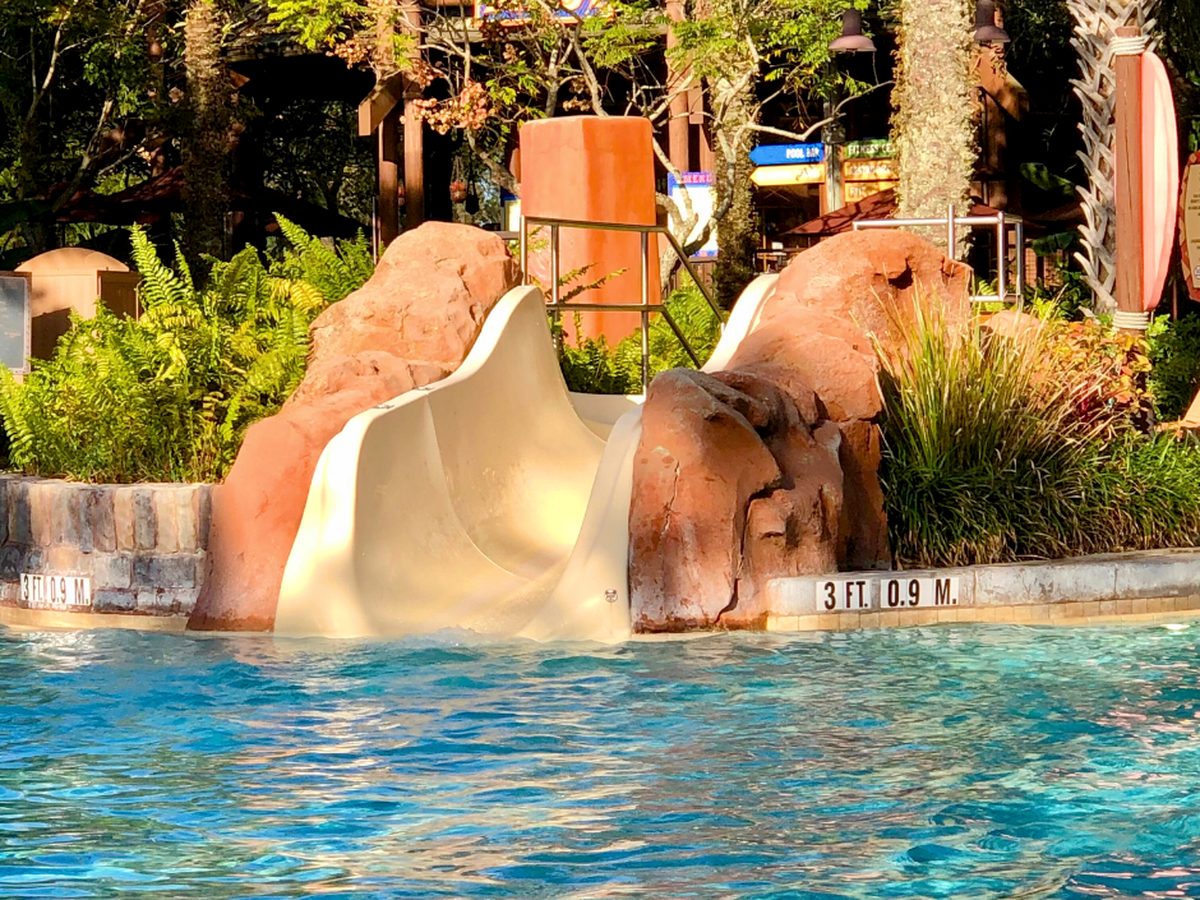 The end of a waterslide between red rockwork at a pool at Disney's Animal Kingdom Lodge, with a 3 ft depth marker on the pool edge and palms and planting behind