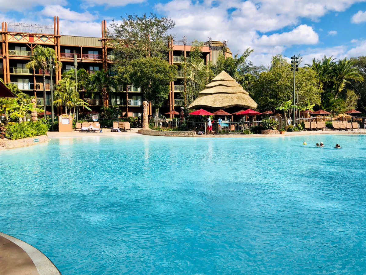 A large turquoise pool at Disney's Animal Kingdom Lodge with a thatched-roof pool bar and red parasols along the far side, and the lodge's balconied guest wings behind