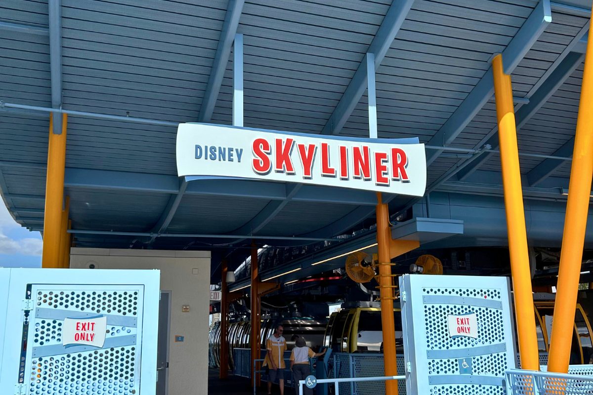The entrance to the Disney Skyliner gondola station, orange support columns under a blue canopy with the red-and-white Disney Skyliner sign and gondolas loading behind
