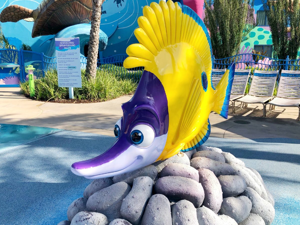 A yellow-and-purple fish figure on a rock at the Schoolyard Sprayground at Art of Animation, with the splash area, its rules sign and poolside chairs around it