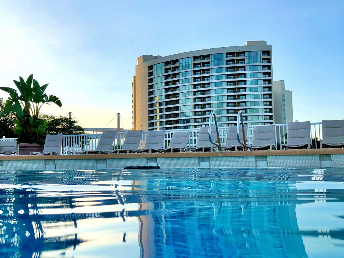 The curved glass face of Bay Lake Tower rising behind a line of sun loungers and a white fence at the edge of a pool at Disney's Contemporary Resort