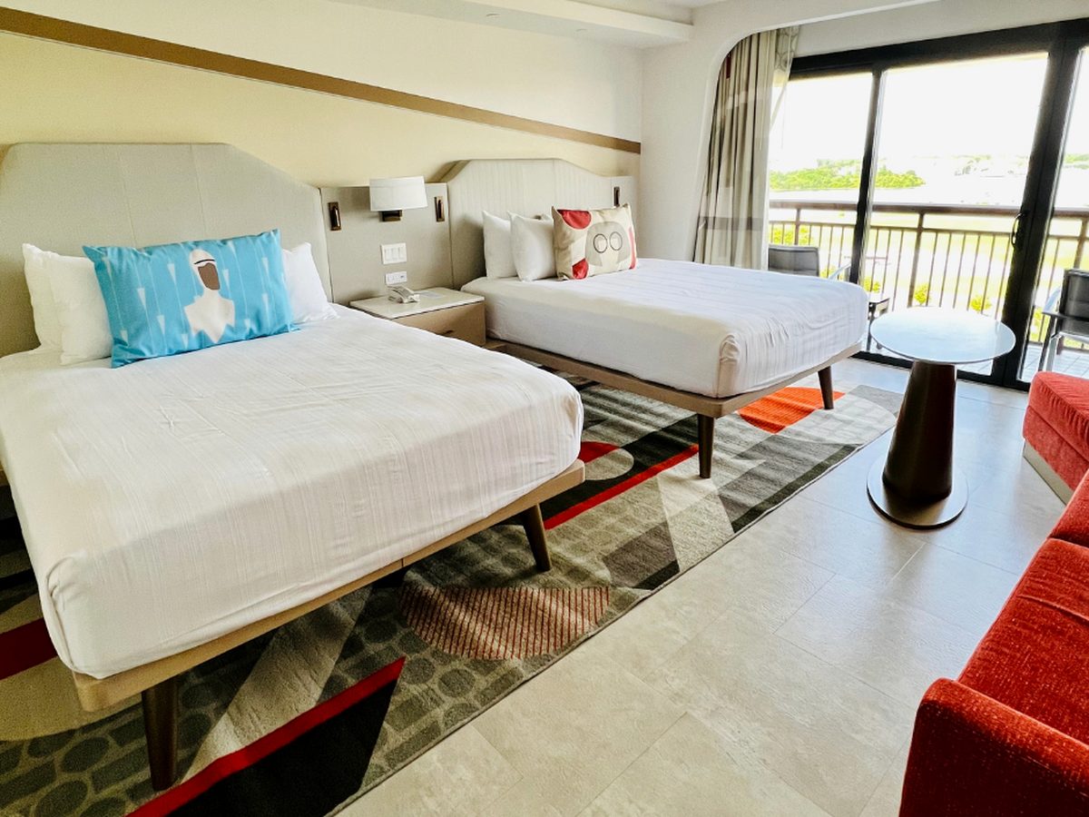 A guest room at Disney's Contemporary Resort with two queen beds, an Elastigirl cushion on one and an Edna Mode cushion on the other, a red sofa and a geometric Incredibles rug, with a balcony beyond