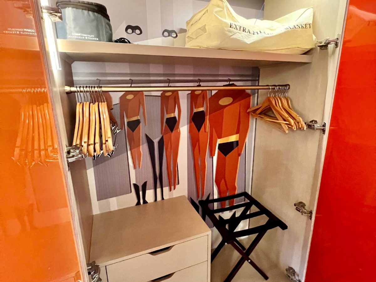 An open guest-room wardrobe at Disney's Contemporary Resort with a row of Incredibles super-suits printed across the back wall, wooden hangers and a luggage rack