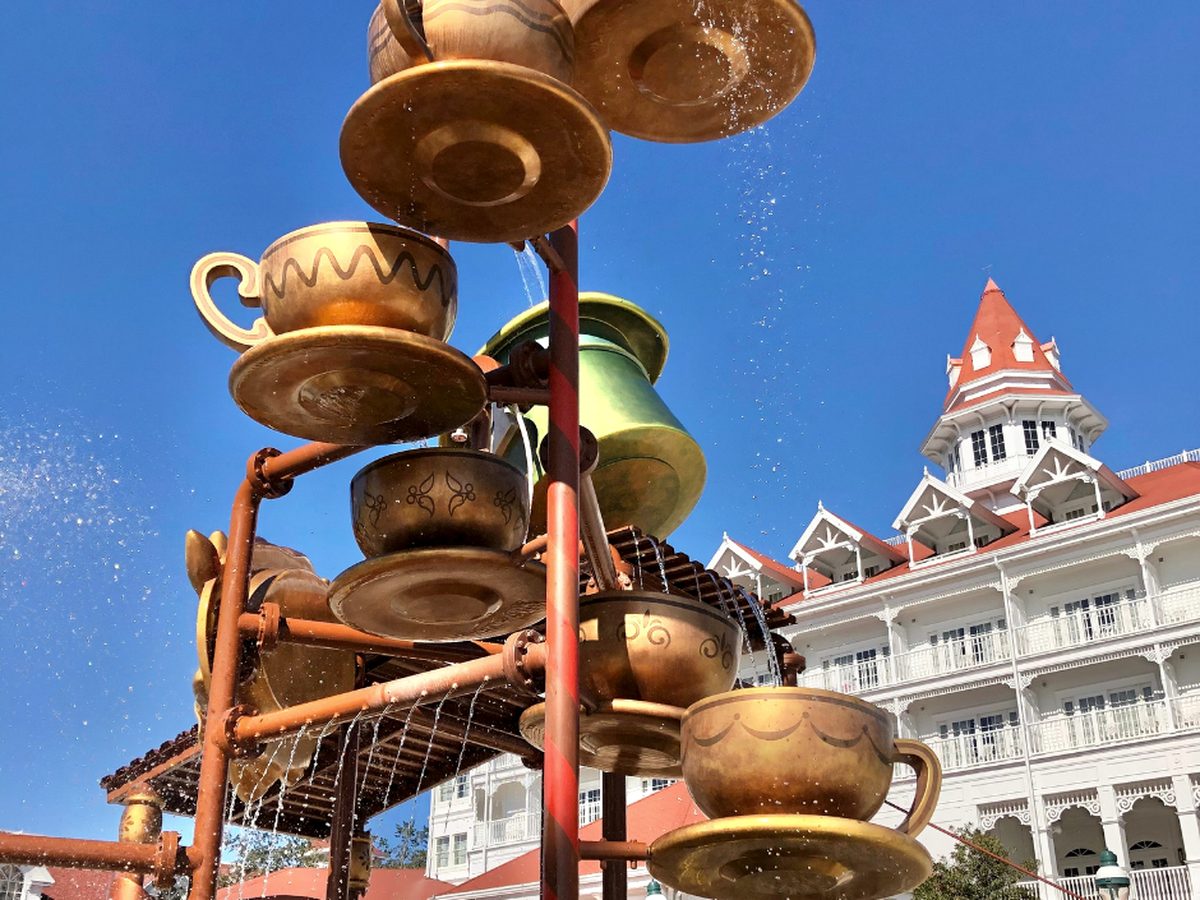 Golden teacup and teapot fountains spilling water in the Alice in Wonderland splash area at Disney's Grand Floridian, with a white resort building and red turret behind