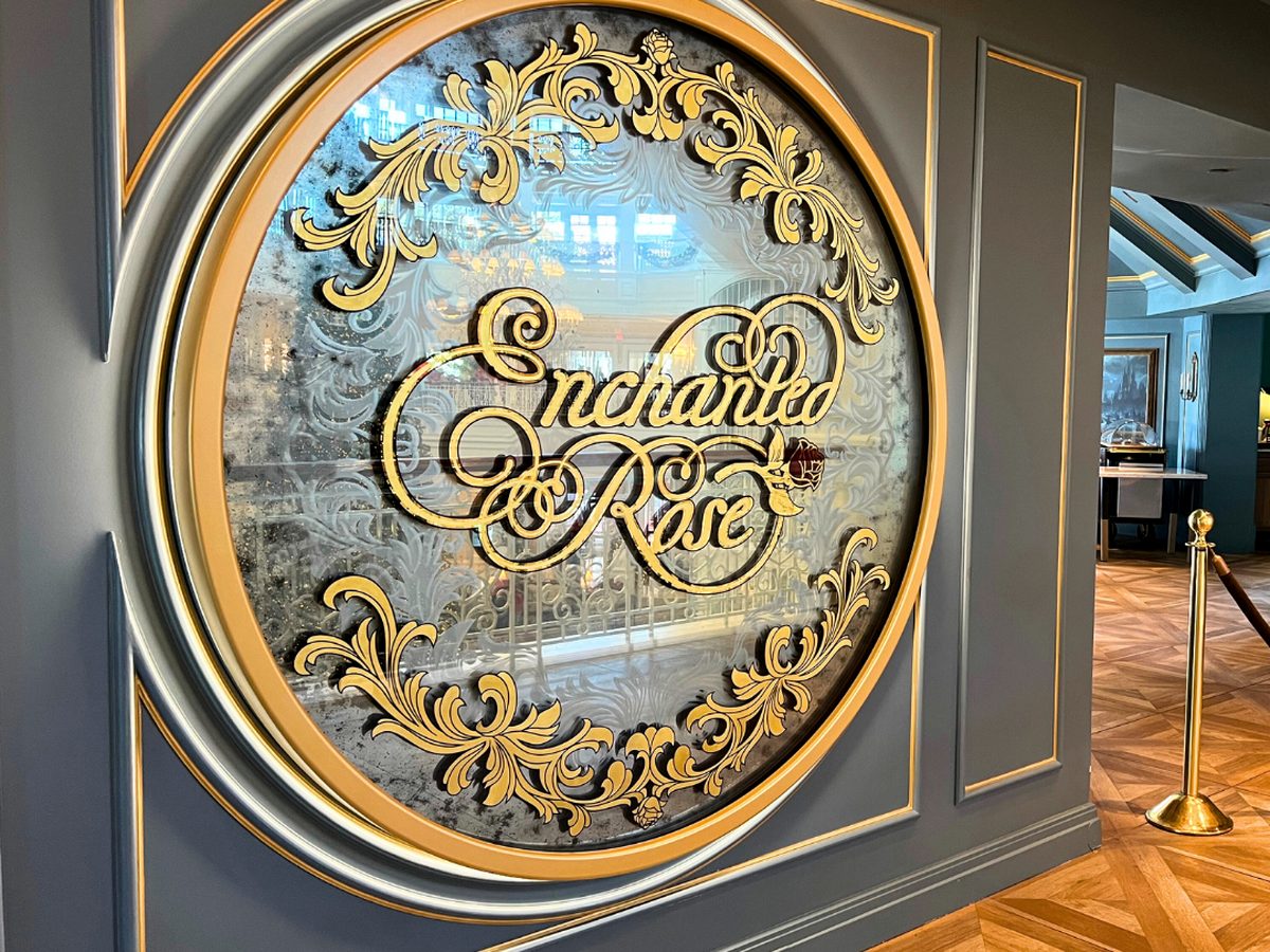 The round mirrored Enchanted Rose sign in gold scrollwork on a grey panelled wall at the entrance to the lounge, with a parquet floor and brass rope barrier beside it