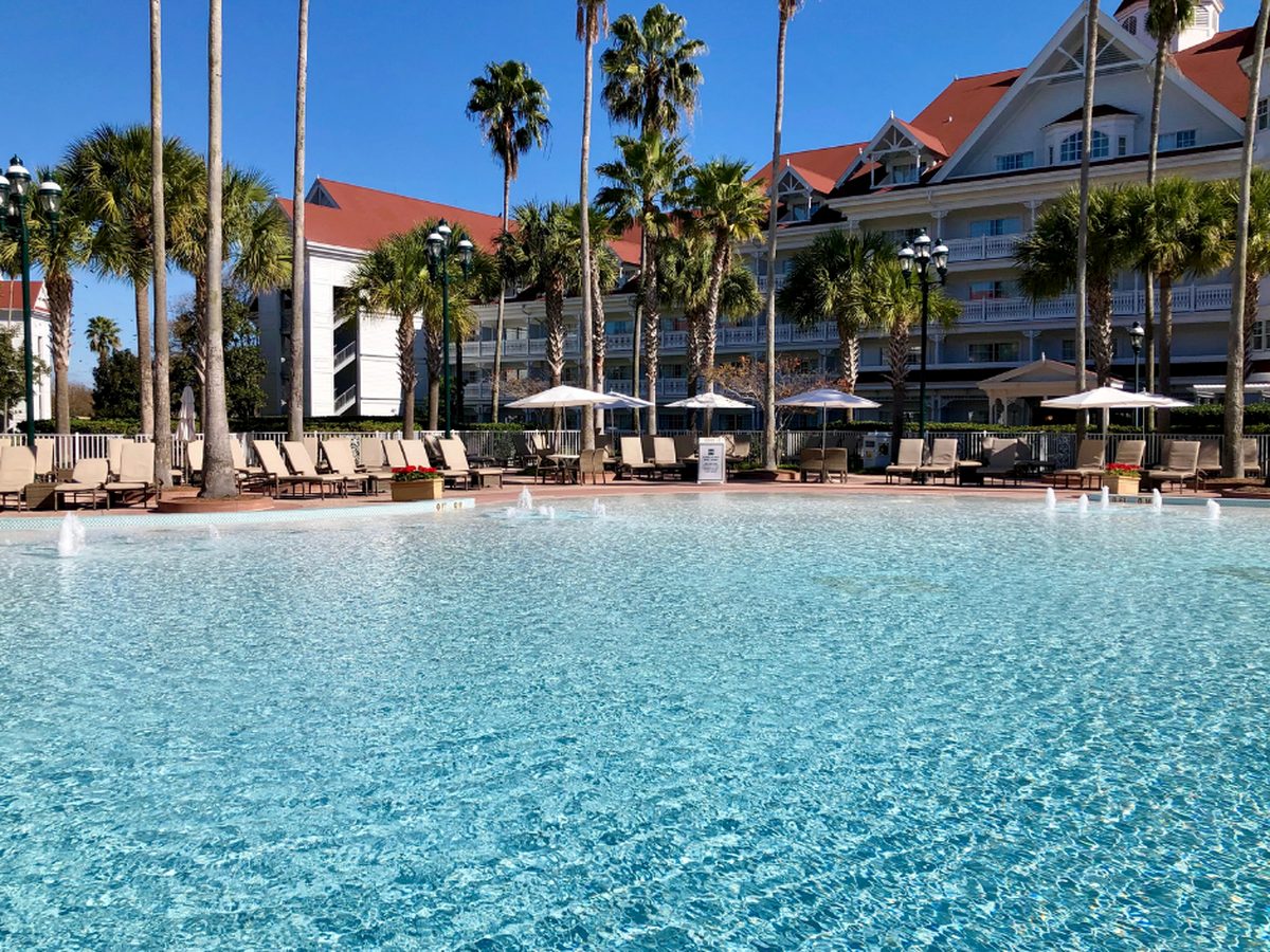 A wide pool at Disney's Grand Floridian with small fountain jets, ringed by sun loungers and parasols, with the white clapboard and red roofs of the resort buildings behind