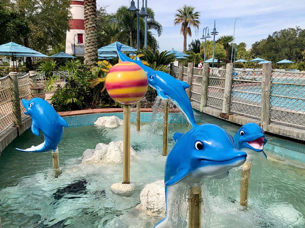 Blue dolphin fountains spouting water in the children's pool at Disney's Old Key West Resort, with the resort's red-and-white lighthouse and pale-blue parasols behind