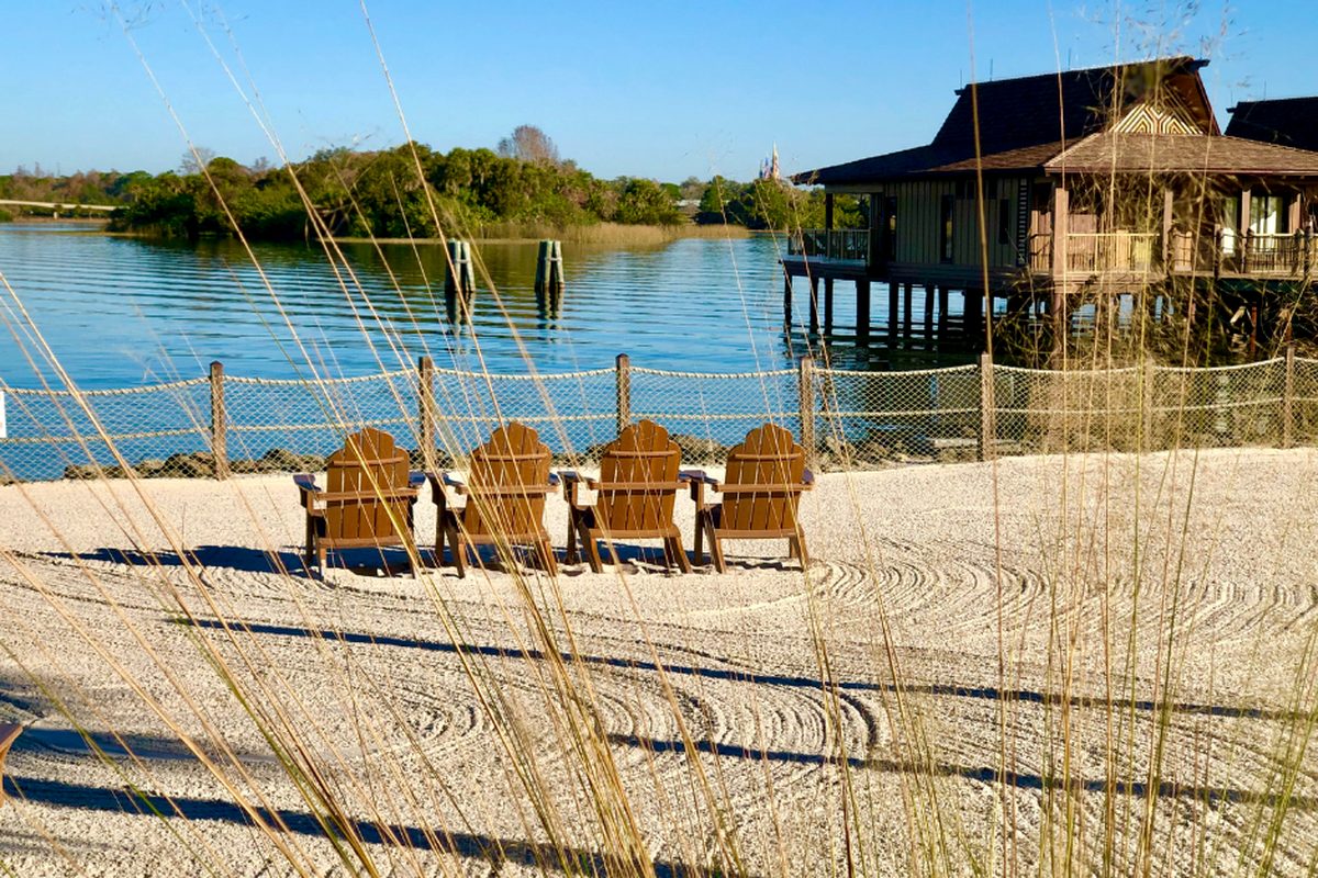 The white-sand beach at Disney's Polynesian Village Resort, four wooden chairs facing the Seven Seas Lagoon with the over-water bungalows on stilts to the right