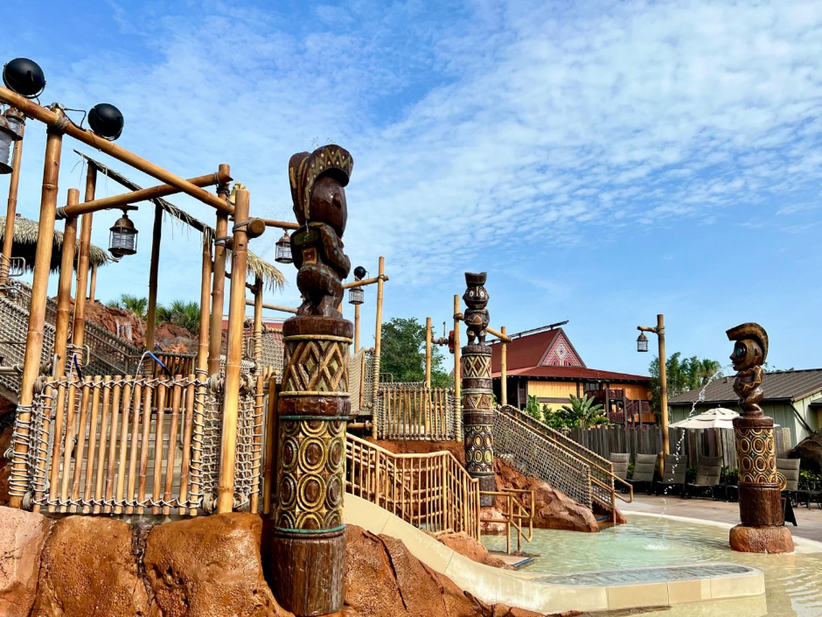 The Kiki Tikis splash area at Disney's Polynesian Village Resort, carved tiki fountains spraying water over a shallow play pool, with a bamboo play frame and a longhouse behind