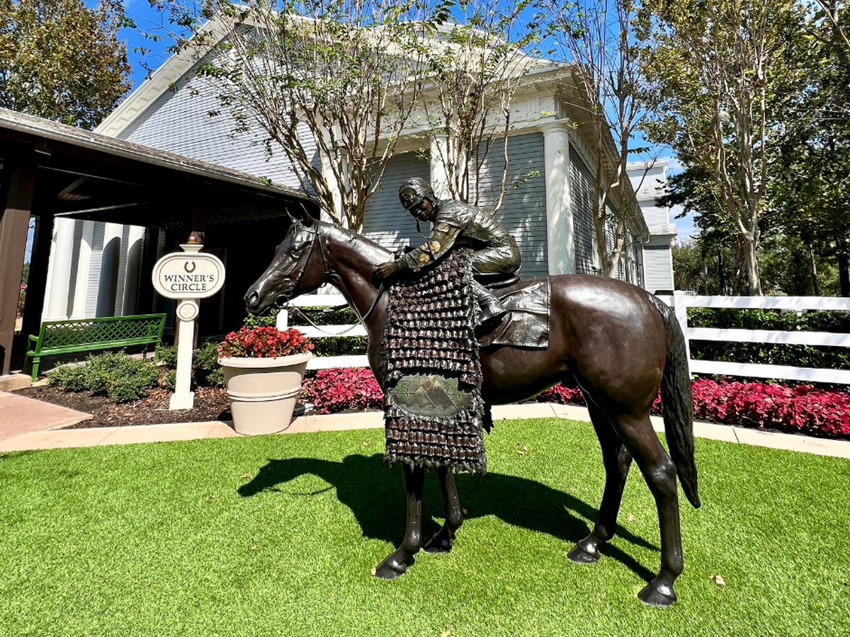 A bronze statue of a racehorse and jockey beside a Winner's Circle sign at Disney's Saratoga Springs Resort, ringed by white fencing and red flowers