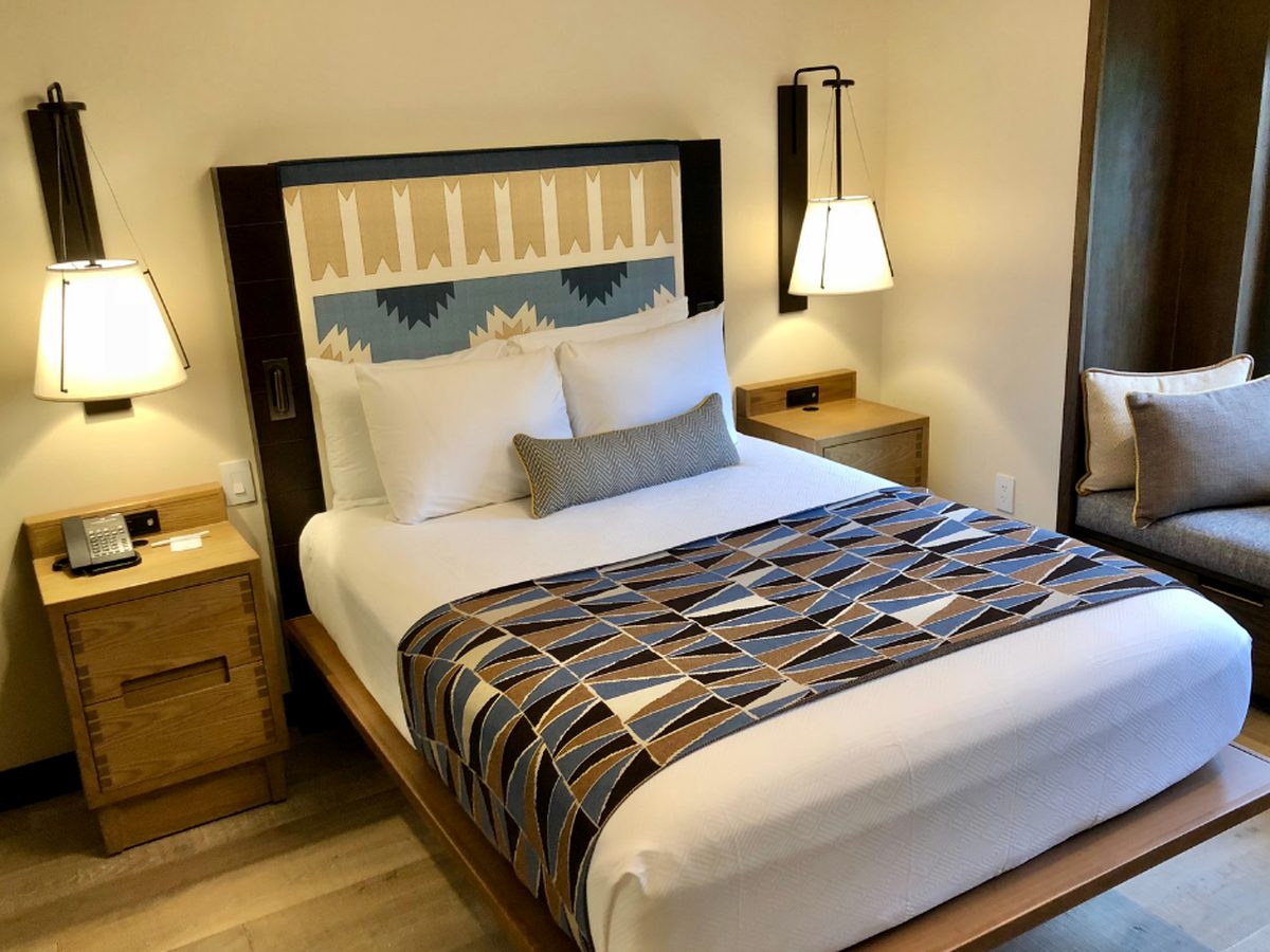 A bedroom in a Cascade Cabin at Disney's Wilderness Lodge, a bed with a geometric blue-and-tan blanket and arrow-motif headboard between hanging lamps, with a window seat beside it