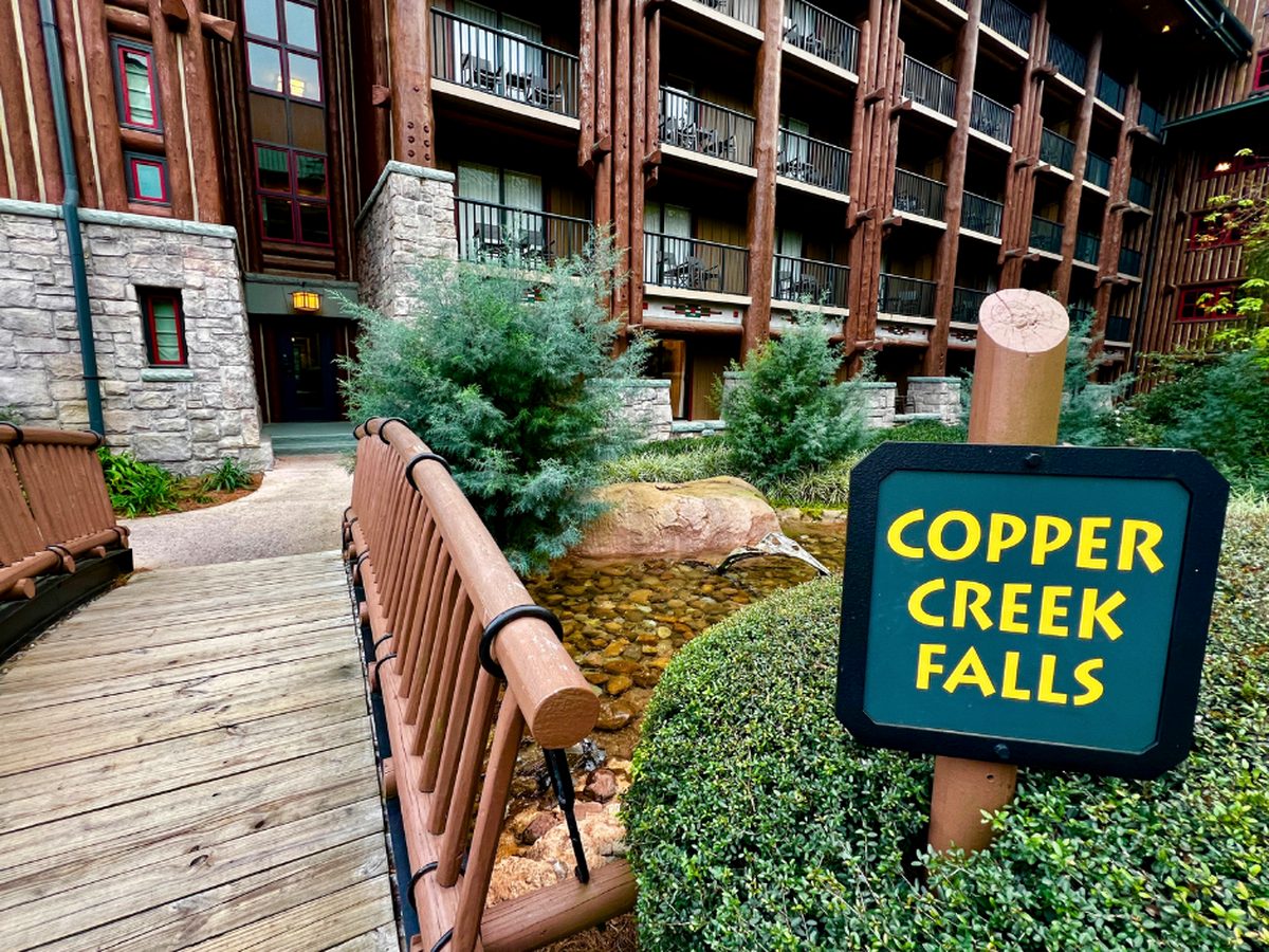 The Copper Creek Falls sign beside a wooden footbridge over the creek in the courtyard at Disney's Wilderness Lodge, with log columns and balconied guest wings behind