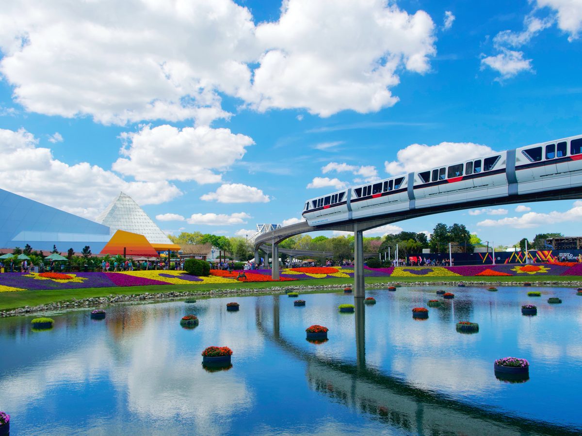 A monorail crossing above festival flower beds and floating flower rafts on the water, with the Imagination pavilion pyramids behind