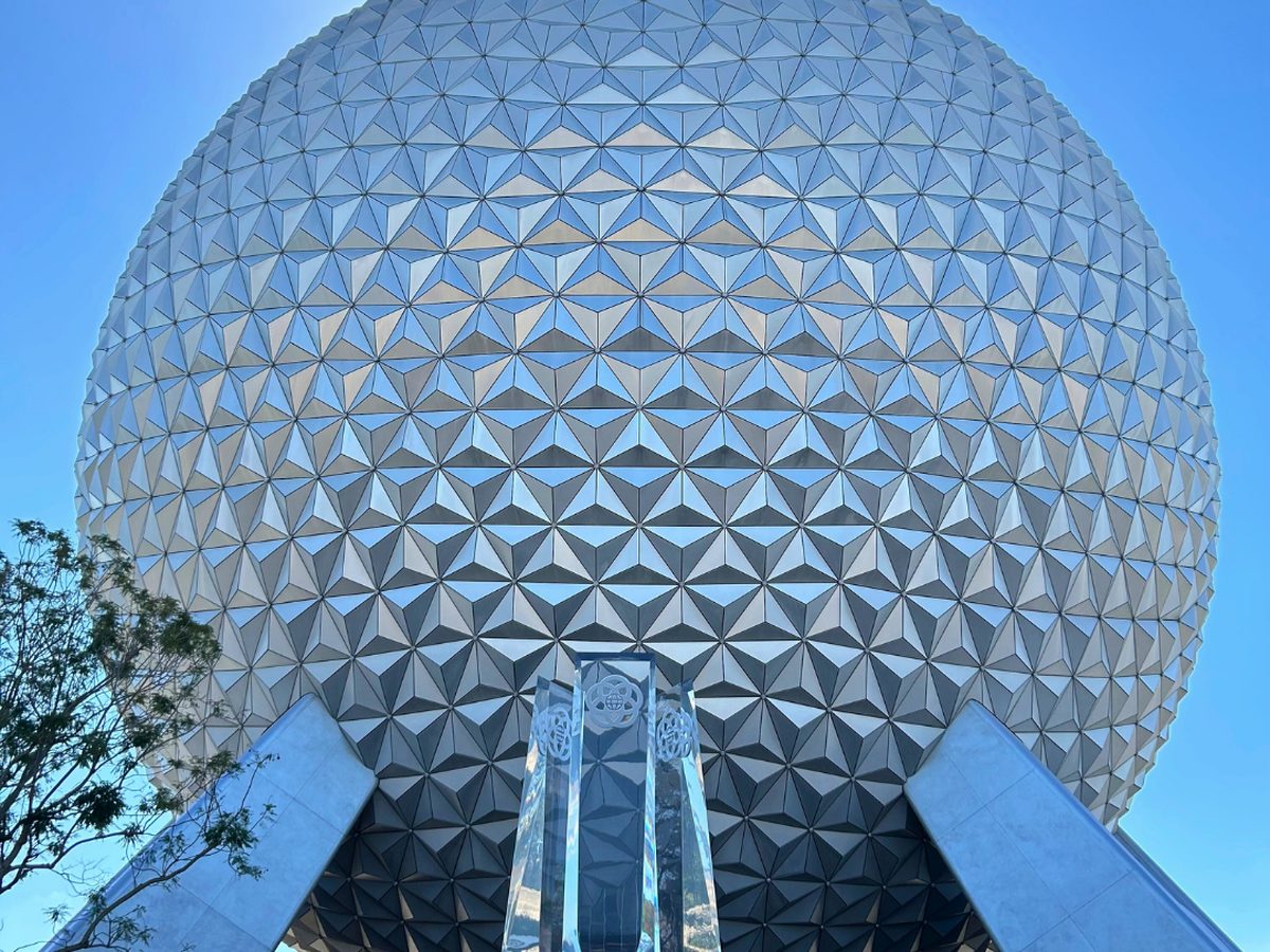Spaceship Earth filling the sky above its silver support legs and the EPCOT-logo pylon at the park entrance