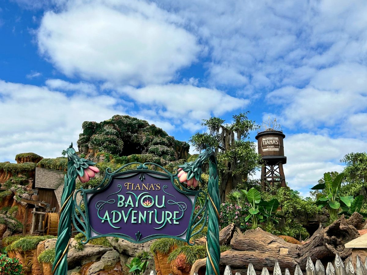 Tiana's Bayou Adventure entrance sign at Magic Kingdom, with the green mountain and Tiana's Foods water tower behind