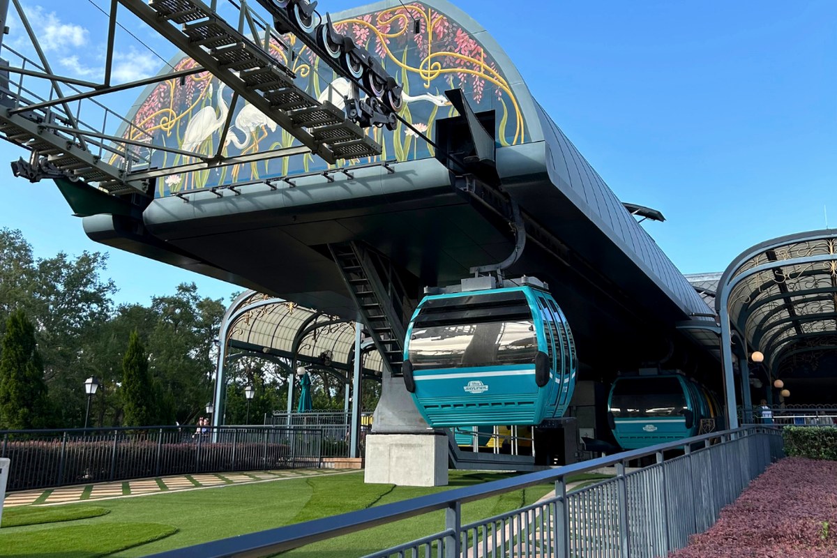 Teal Skyliner gondolas arriving under the decorated canopy at EPCOT's International Gateway station, against a clear blue sky