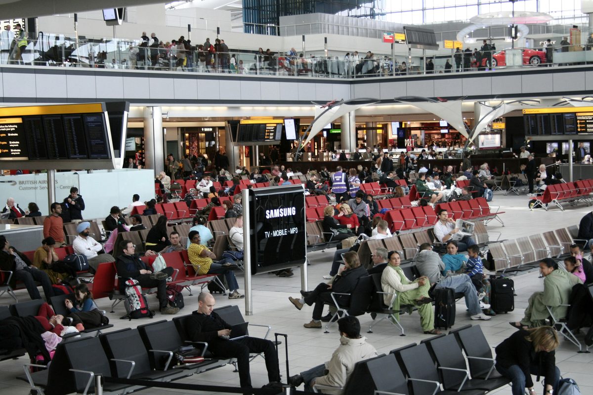 Travellers seated with bags beneath departure boards in a Heathrow terminal.