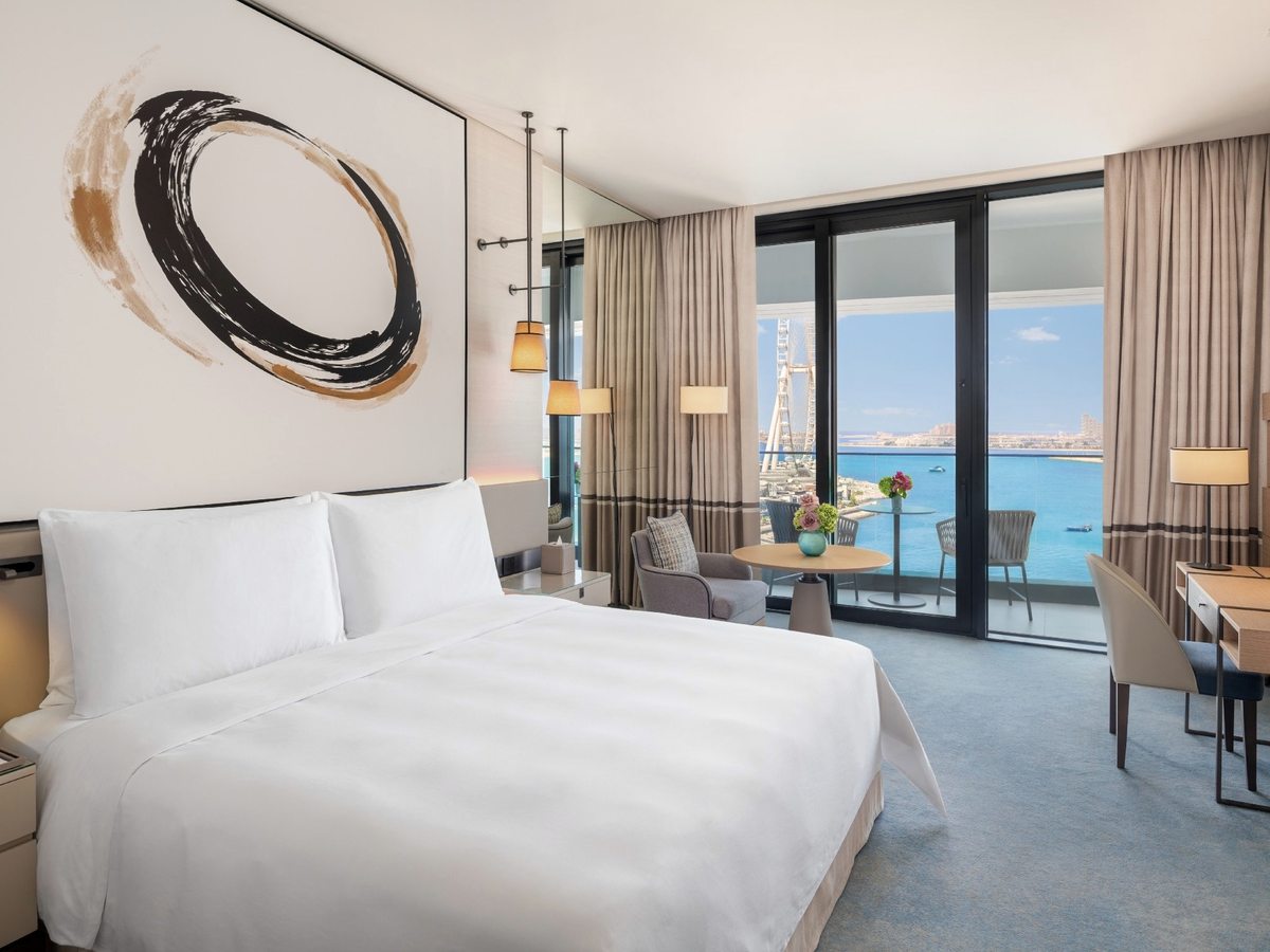 A hotel room with a king bed dressed in white, a circular brushstroke artwork above the headboard and floor-to-ceiling windows to a seating area and the sea.