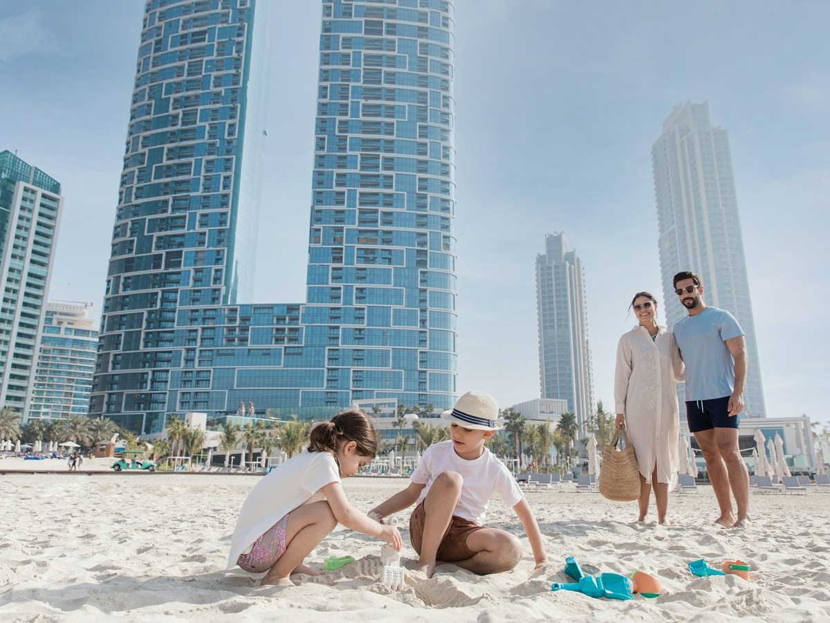 Two children building sandcastles on the soft sand, with the tall JBR towers behind them in bright sun.