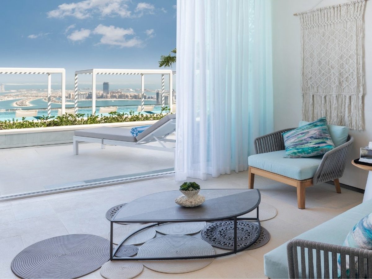 A room opening through glass doors onto a private balcony furnished with a daybed, sofa and low table, with the city beyond.