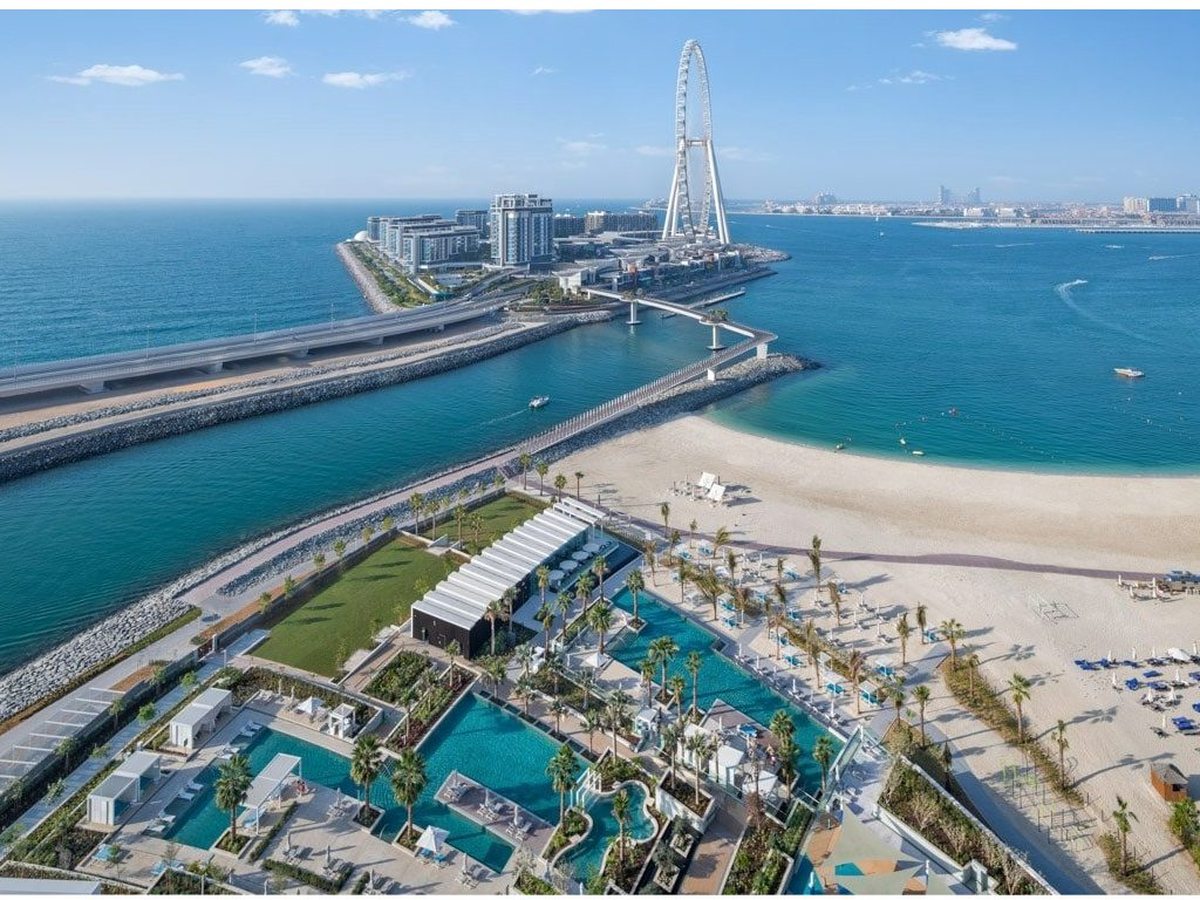 An aerial view of the resort's terraced pool gardens and reserved sandy beach beside turquoise sea, with a footbridge to Bluewaters Island and the Ain Dubai wheel.