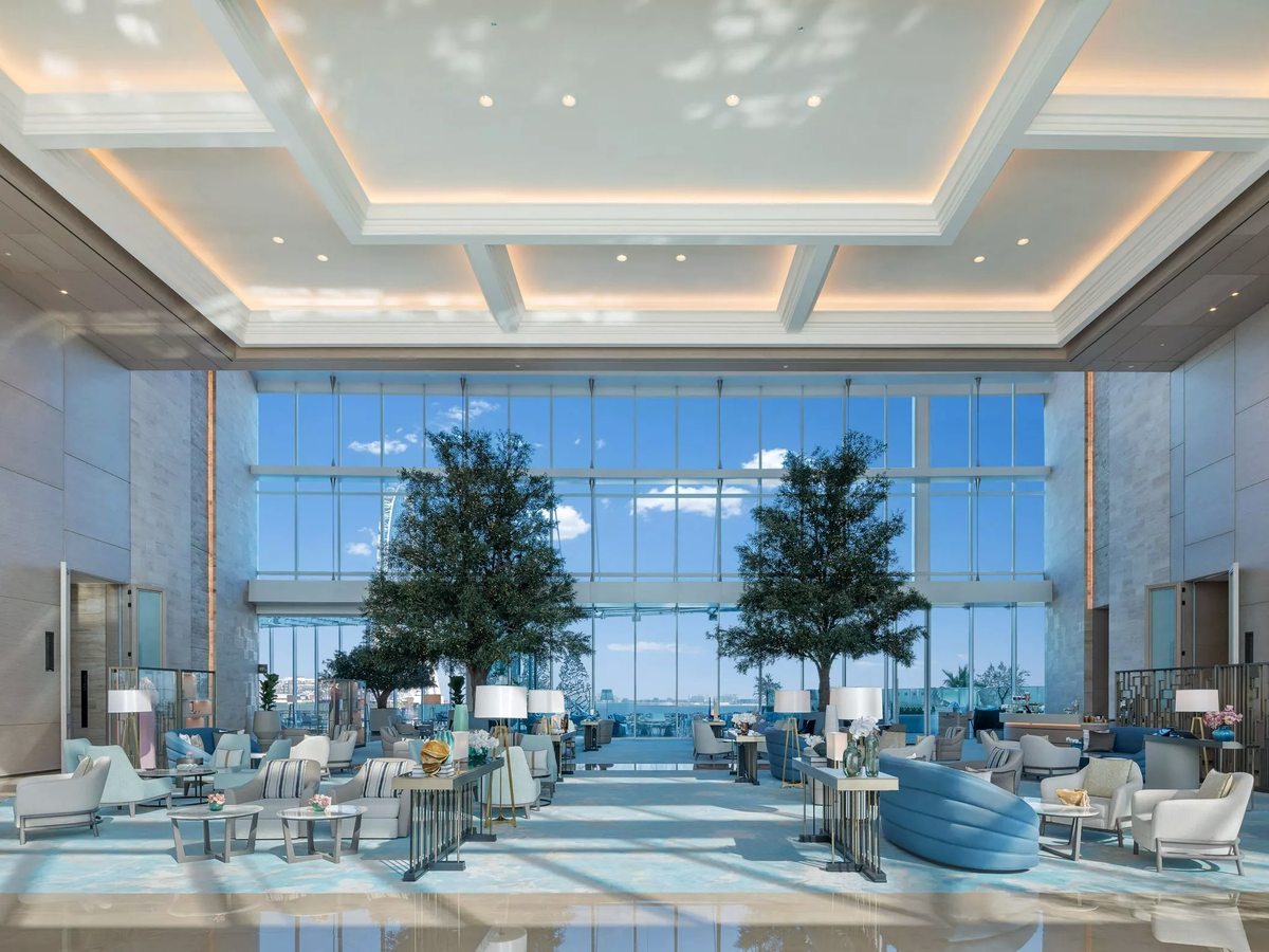 A double-height lobby lounge with pale blue and cream sofas and armchairs, indoor trees, a coffered ceiling and full-height windows to the sea.