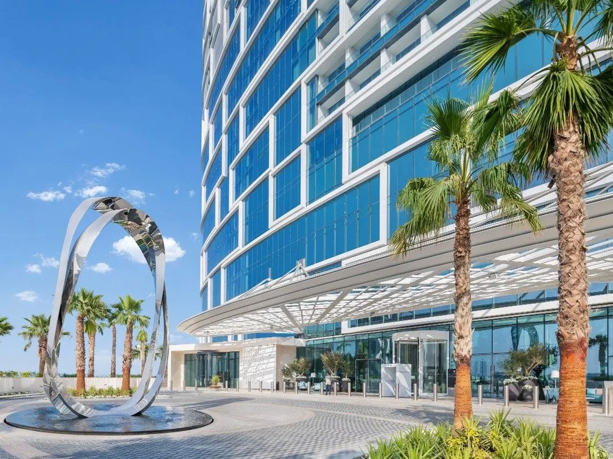The landscaped base of the towers with tall palms, a large polished chrome ring sculpture and paved walkways in bright sun.