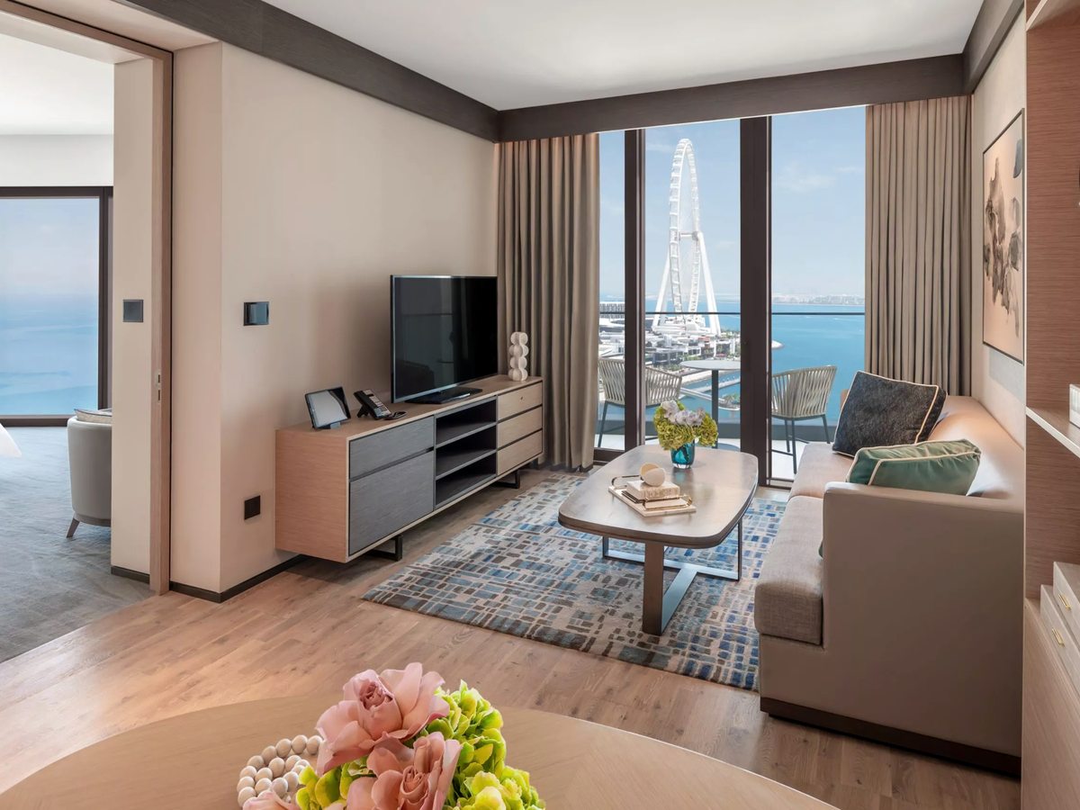 A suite living room with a grey sofa, coffee table and media console, floor-to-ceiling windows framing Ain Dubai and the sea, and the bedroom through an open doorway.