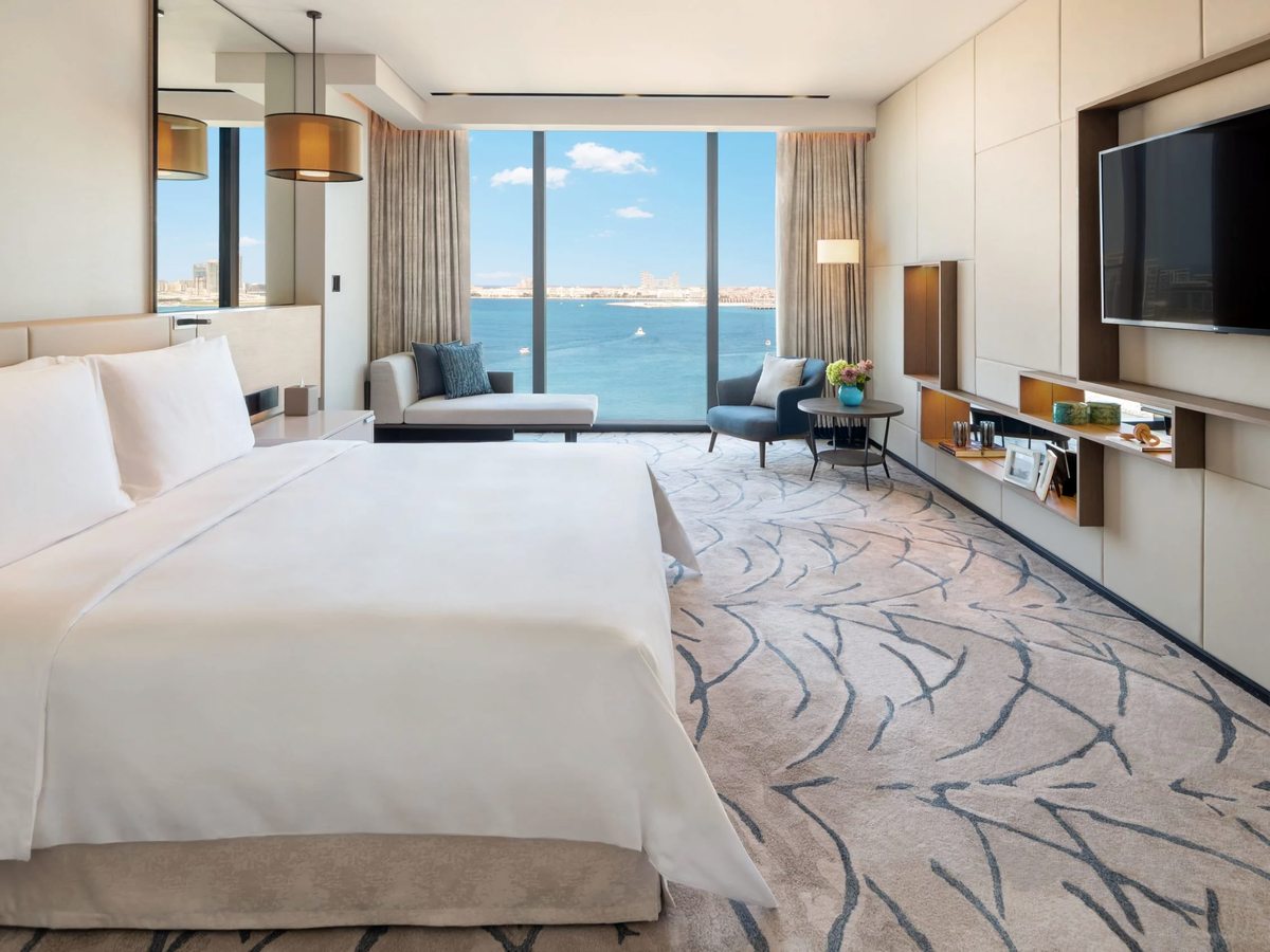 A suite bedroom with a king bed, a wave-patterned blue carpet, a wall-mounted television and full-height windows over the sea.
