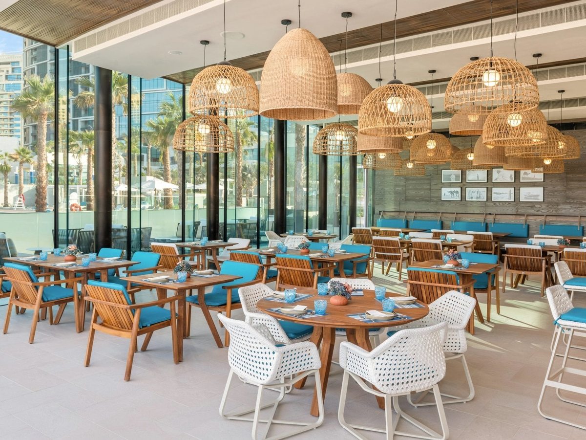 A casual restaurant under woven rattan pendant lamps, with wooden tables and blue chairs and glass walls opening to the pool and palms.
