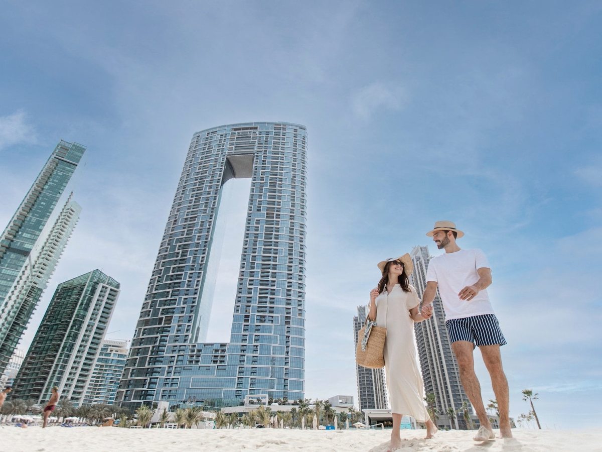 The two glass towers rising above The Walk, a palm-lined seafront promenade, with people strolling in bright sun.