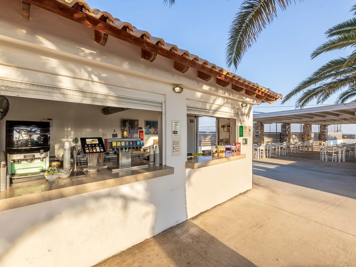 An open beach bar kiosk with a serving counter, drinks fridges and shaded seating under straw parasols by the sea.