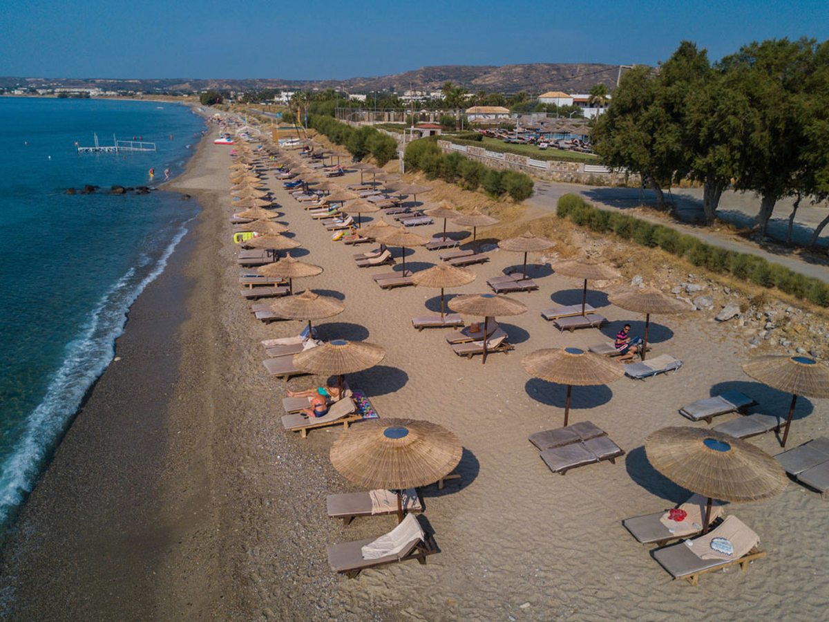 An elevated view of a long organised beach lined with straw parasols and sun loungers, with a coastal path behind.