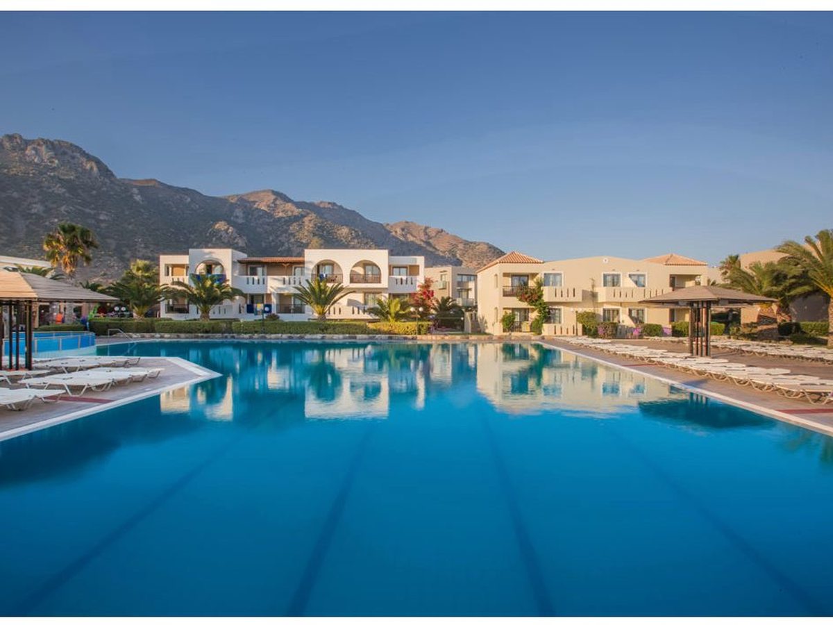 A large rectangular freshwater pool edged with sun loungers and thatched parasols, accommodation blocks and hills behind.