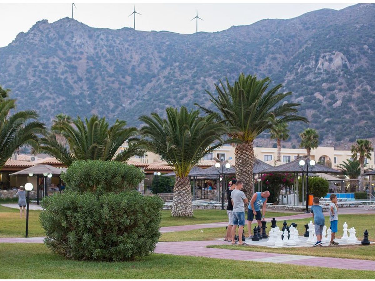 A giant outdoor chess set on a paved square in the lawned gardens, with palms, low white buildings and hills behind.